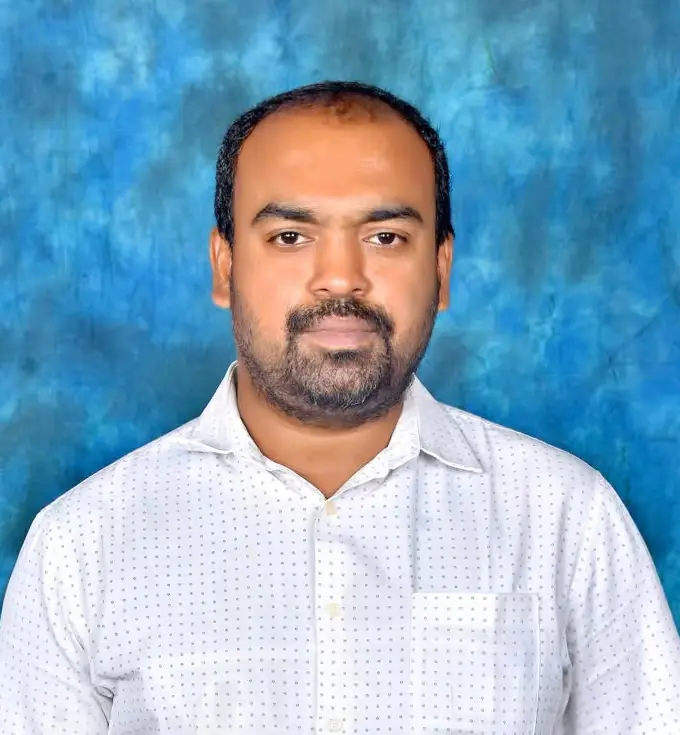 A man with short hair and a beard is wearing a white, patterned shirt. He stands against a blue textured background.