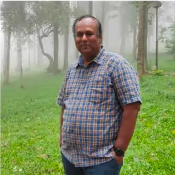 Man in a checkered shirt stands in a foggy, wooded area with grass underfoot.
