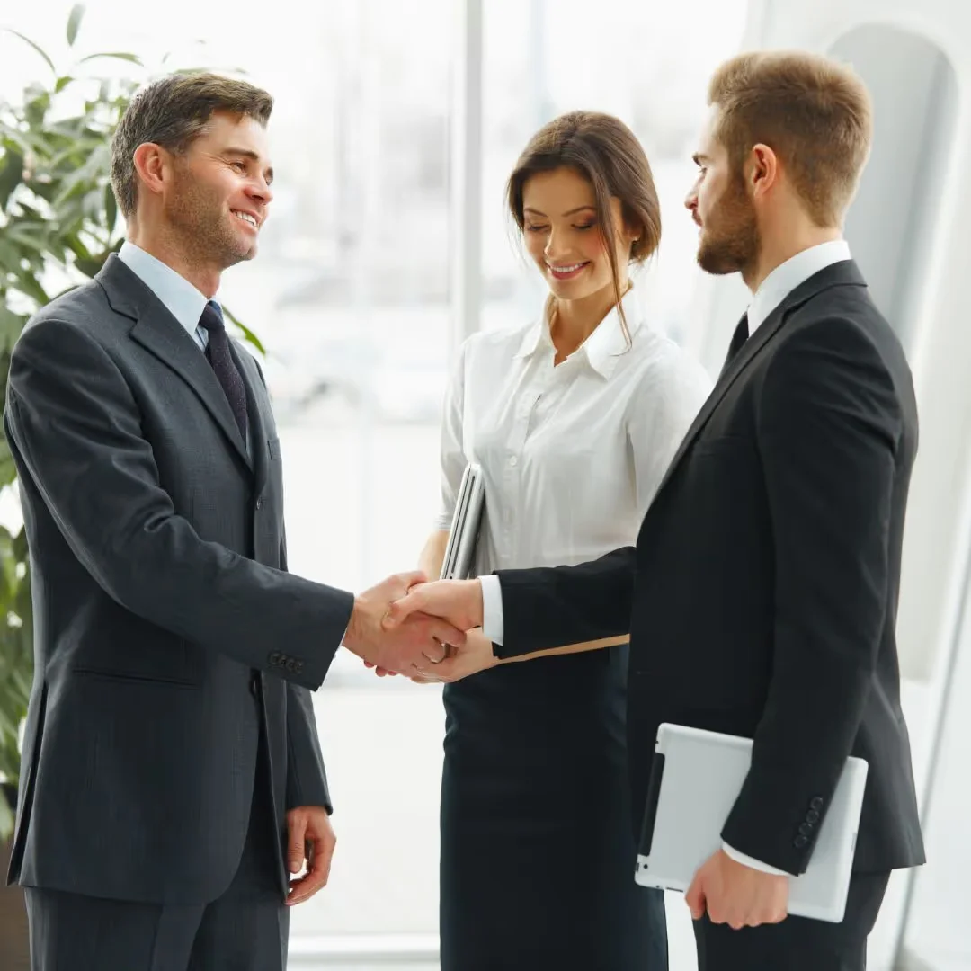 Three business professionals in formal attire converse; two men are shaking hands, while a woman stands between them, holding a tablet.